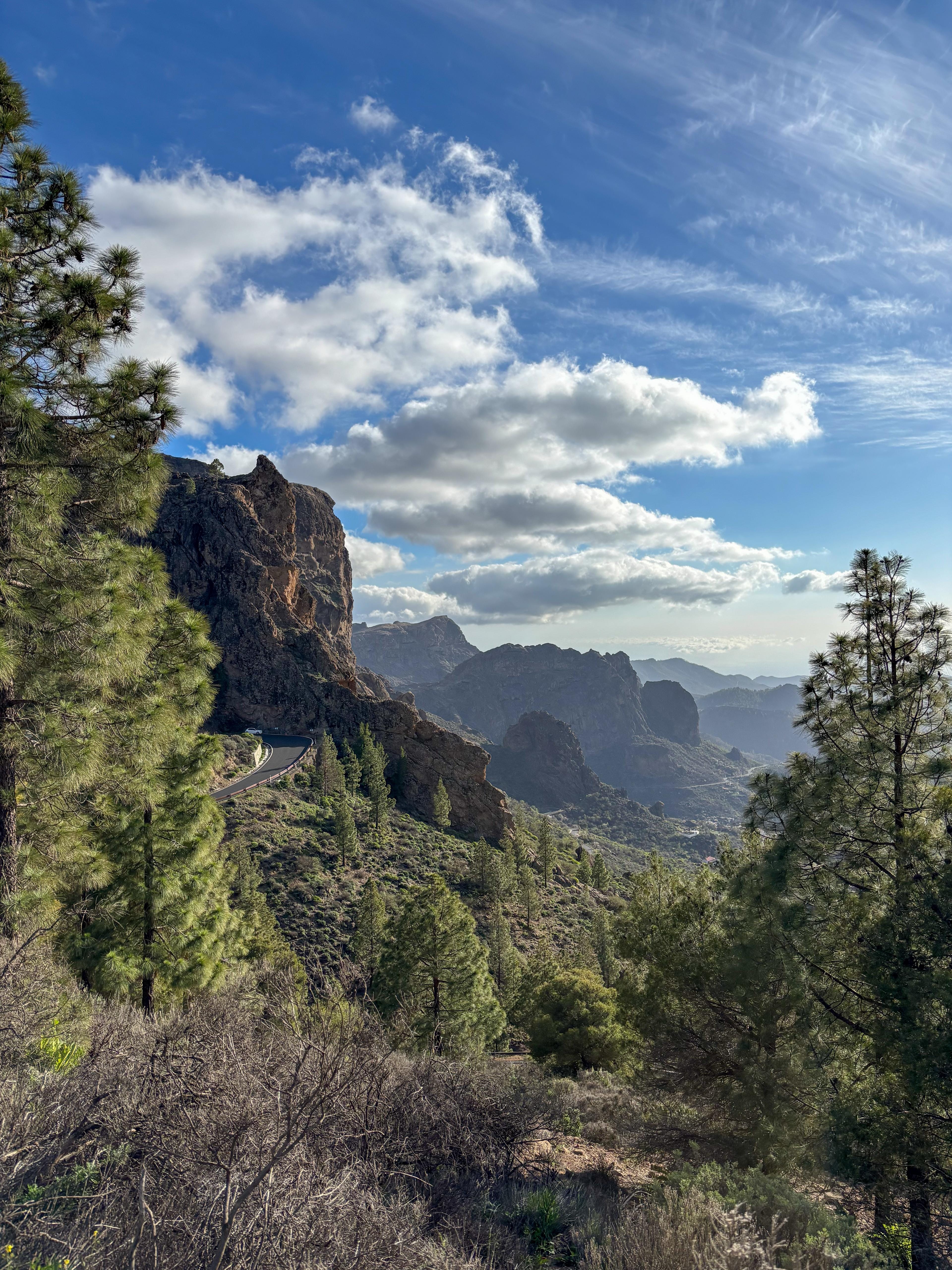 Panoramic view from Roque Nublo trail showing dramatic volcanic rock formations, pine forests, and layered mountain ranges of central Gran Canaria under a bright blue sky with scattered clouds. A winding mountain road can be seen cutting through the rugged landscape